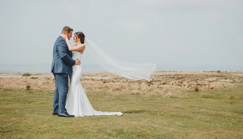 Bride and groom embrace in an open field during their wedding day, with her veil flowing in the wind.