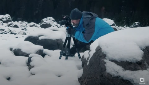 Photographer in blue jacket operating camera on tripod in snowy rocky landscape during winter.