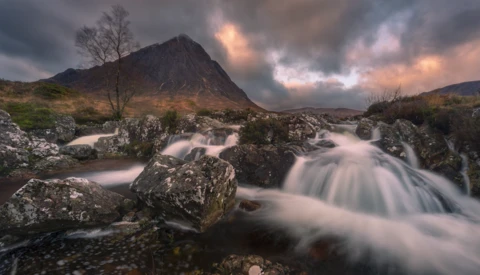 Rushing mountain stream flowing around moss-covered rocks beneath a dramatic peak at sunset.