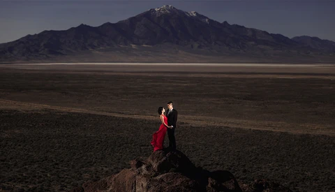 Couple in formal attire standing on a dark rock formation in an expansive desert plain, with a snow-capped volcanic mountain in the distance.