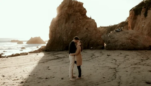 Couple kissing on a sandy beach with dramatic rock formations and ocean waves in the background.