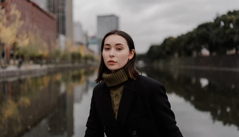 Woman in black blazer and olive turtleneck standing by a calm waterway with urban buildings behind her.
