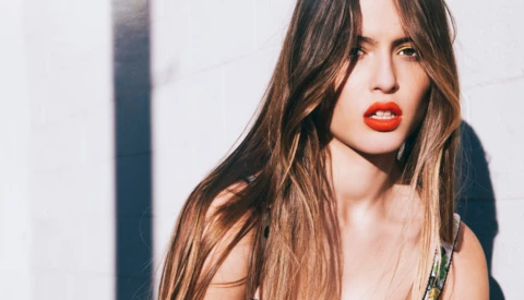 Woman with long brunette hair and red lipstick against a white wall with dramatic shadow.