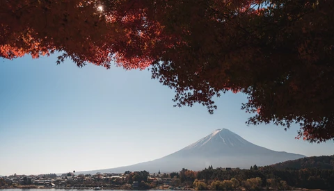 Snow-capped volcanic mountain framed by red autumn foliage in foreground, with clear blue sky and lakeside town below.