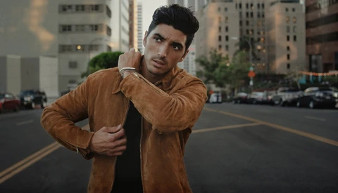 Man in tan suede jacket posing on urban street with city buildings behind.