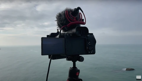 Mirrorless camera mounted on tripod with shotgun microphone against overcast ocean horizon.