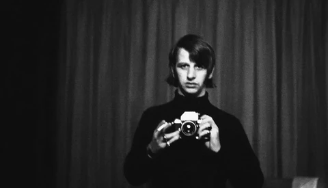 Man in dark sweater holding vintage camera directly at lens against curtained backdrop.