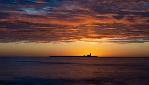 Silhouetted lighthouse on a distant island at sunset over calm ocean waters.
