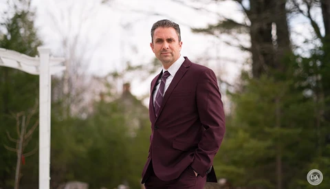 Man in burgundy suit standing outdoors with evergreen trees in background.