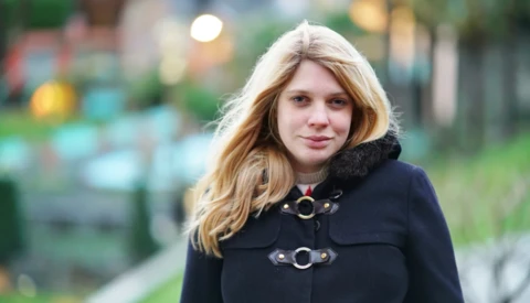 Woman with long blonde hair wearing a black coat with decorative buckles, photographed outdoors with blurred background.