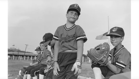 Baseball players in vintage uniforms stand on a field with stadium structures visible in the background.