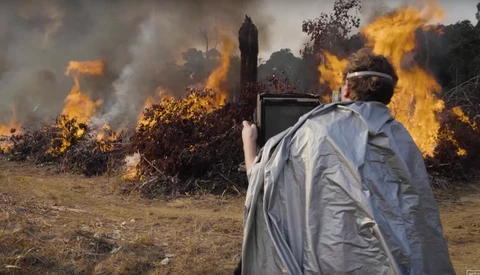 Person in light-colored clothing operating equipment while managing large outdoor fire with intense flames.