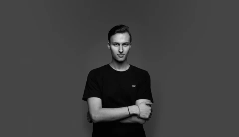 Young man in black t-shirt with arms crossed, looking at camera against gray background.