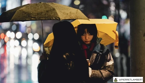 Two people under yellow umbrellas at night on a wet street with blurred city lights in the background.