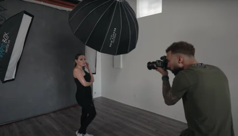 Photographer with camera on tripod shooting portrait of woman in black clothing against gray backdrop with studio lighting setup.