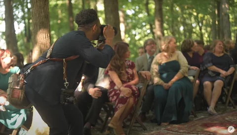 Photographer with camera on shoulder capturing an outdoor forest ceremony with seated guests in background.