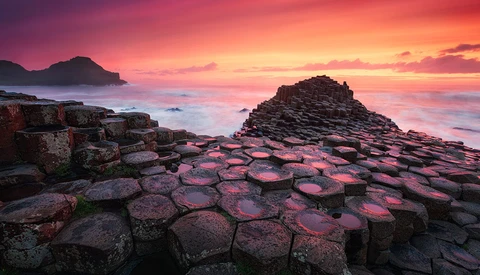 Hexagonal basalt columns in foreground with golden sunset sky and rocky islands in background.