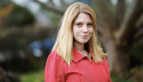 Blonde woman in coral pink shirt smiling outdoors with blurred greenery background.