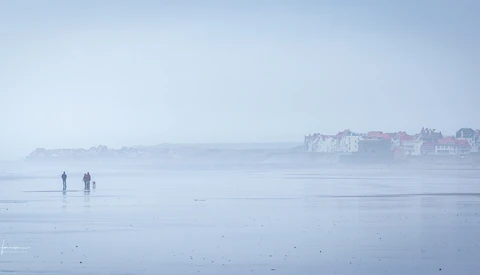 Two figures walking on a misty, wet beach at dawn with a coastal town silhouetted in the distance.