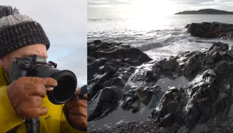 Photographer in yellow jacket and knit beanie holding telephoto lens, with coastal rocky shoreline scene in background.