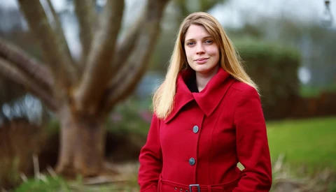 Young woman with long blonde hair wearing a bright red coat, photographed outdoors with soft natural lighting.
