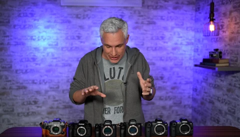 Man demonstrating six camera bodies arranged in a row on a table with purple brick wall backdrop.