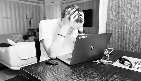 Person in white shirt holding head while working at laptop in hotel room.