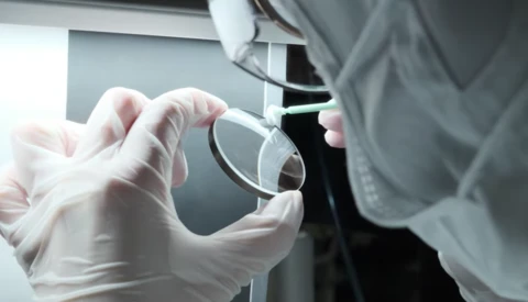Gloved hands holding a clear glass lens or optical element during laboratory examination.