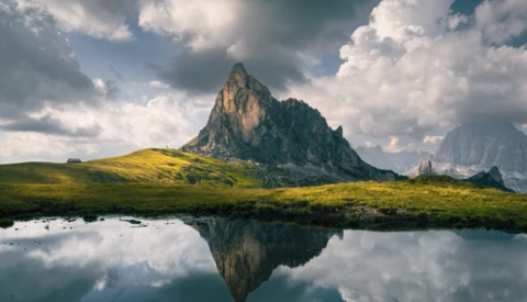 Dramatic alpine peak reflected in still water, surrounded by green meadow and dramatic cloudy sky.