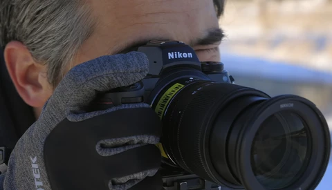 Photographer holding a Nikon DSLR camera with telephoto lens, wearing gray gloves and winter clothing.