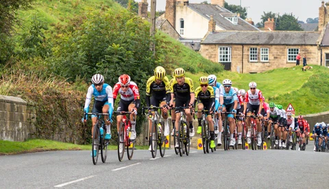 Professional cycling race with peloton riding on rural road past stone buildings and green hillsides.
