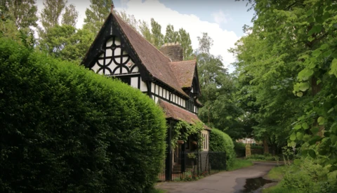 Half-timbered Tudor-style cottage surrounded by lush green hedges and trees.