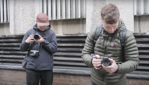 Two photographers holding compact cameras outside a modern building with corrugated metal siding.