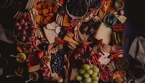 Overhead flat lay of cured meats, cheeses, fruits, and appetizers arranged on a dark surface with scattered forks.