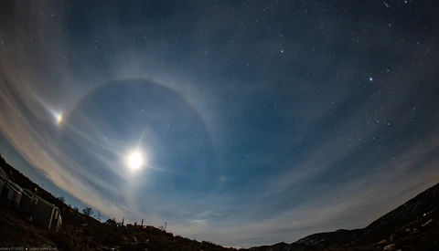 Bright moon with concentric halo rings in twilight sky above dark mountain silhouettes.