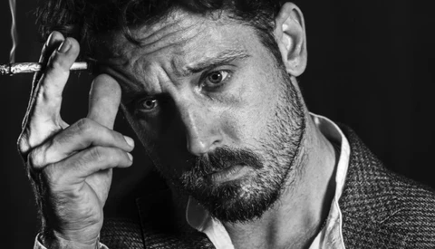 Bearded man in black and white portrait holding cigarette near his face.