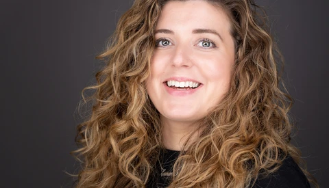 Woman with long wavy brown hair smiling at camera against dark background.