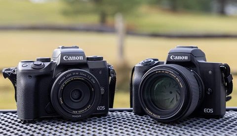 Two Canon EOS mirrorless cameras side by side on a checkered surface with blurred green background.