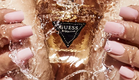 Hands with pink nail polish holding a GUESS fragrance bottle submerged in water.