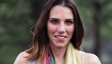 Woman with shoulder-length brown hair smiling at camera outdoors with blurred green foliage background.