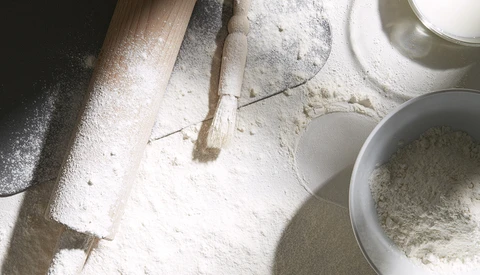 Overhead view of baking ingredients including flour, a wooden rolling pin, pastry brush, and bowls on a white surface.