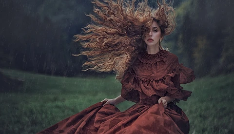 Woman in burgundy dress with windswept long hair in heavy rain against dark green field.