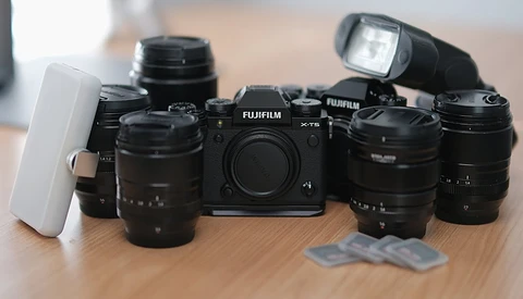 Fujifilm X-T5 camera body surrounded by multiple black lenses, a white diffuser, and external flash on wooden surface.