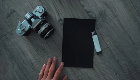 Flat lay of vintage rangefinder camera with lens, black notebook, and photographer's hand on wooden surface.