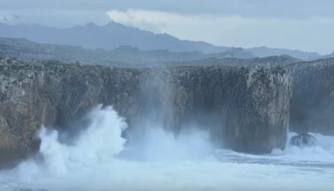 Massive waves crash against basalt columnar cliffs under stormy skies with mountains in the distance.