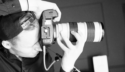 Photographer holding a mirrorless camera with a telephoto lens to their face while shooting.