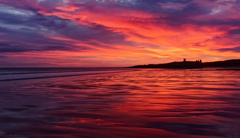 Vibrant sunset over calm ocean water with silhouetted headland and structures on the horizon.