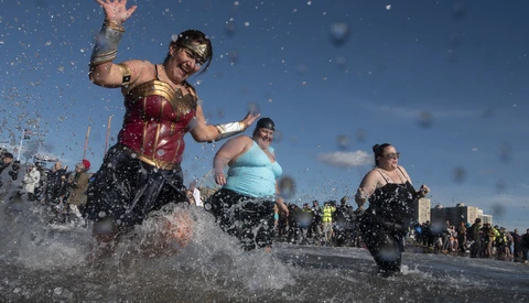 Multiple people running through shallow water on a beach under blue sky with water splashing around them.
