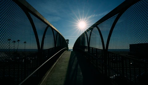 Silhouetted pedestrian bridge with mesh railings framing a bright sun against blue sky.