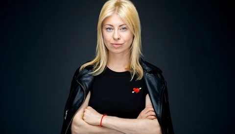Blonde woman in black leather jacket and black shirt against dark background, arms crossed.